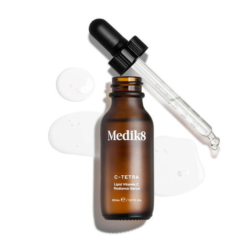 Záber zhora na 30 ml fľaštičku séra Medik8 C-Tetra Lipid Vitamin C Radiance Serum. Jantárová sklenená fľaštička je otvorená a stojí uprostred záberu, pričom jej čierne viečko s kvapkadlom leží šikmo za ňou. Číre tekuté sérum je umelecky rozstreknuté a rozliate na čistom bielom pozadí okolo fľaštičky, pričom na sklenenej pipete je vidieť niekoľko kvapiek. Osvetlenie je jasné, vrhá jemné tiene doľava a vytvára klinickú, no zároveň elegantnú estetiku.