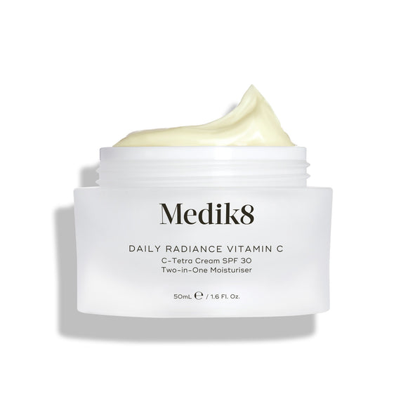 Sklenený kelímok Medik8 Daily Radiance Vitamin C je zobrazený otvorený s bohatou kopou svetložltého krému na vrchu. Text označuje produkt ako 50 ml C-Tetra Cream SPF 30 2-v-1 hydratačný krém.
-7