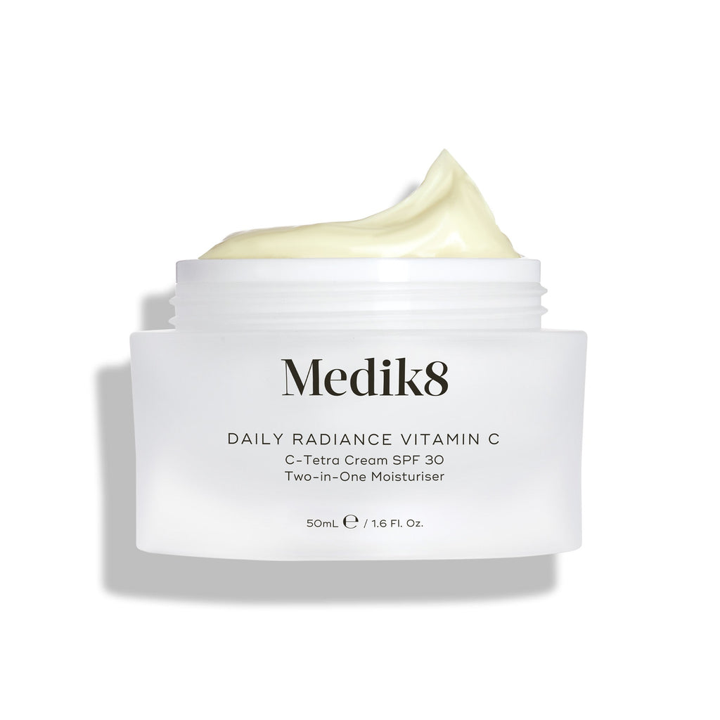Sklenený kelímok Medik8 Daily Radiance Vitamin C je zobrazený otvorený s bohatou kopou svetložltého krému na vrchu. Text označuje produkt ako 50 ml C-Tetra Cream SPF 30 2-v-1 hydratačný krém.
