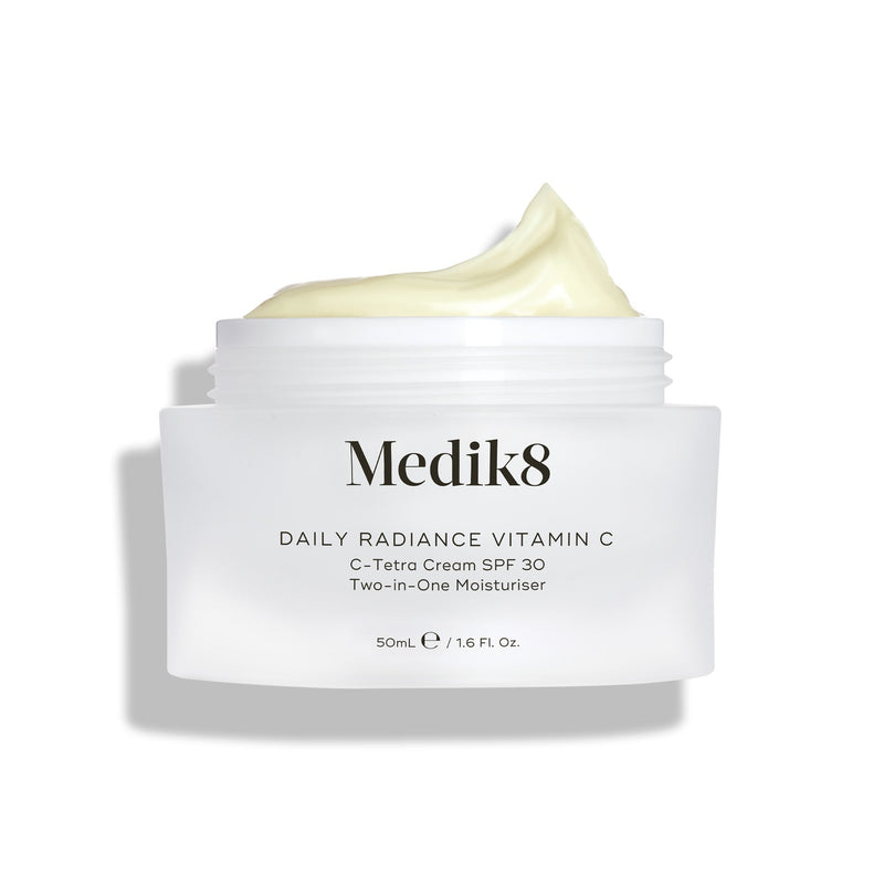 Sklenený kelímok Medik8 Daily Radiance Vitamin C je zobrazený otvorený s bohatou kopou svetložltého krému na vrchu. Text označuje produkt ako 50 ml C-Tetra Cream SPF 30 2-v-1 hydratačný krém.
