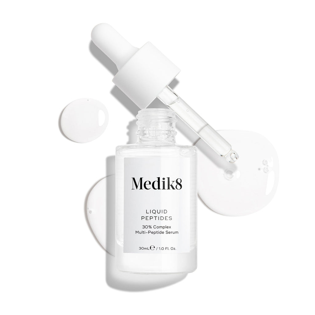 Priehľadná sklenená fľaštička séra Liquid Peptides je zobrazená s vybratým kvapkadlom, ktoré leží vedľa nej spolu s kvapkami séra, ktoré detailne ukazujú jeho tekutú textúru.
