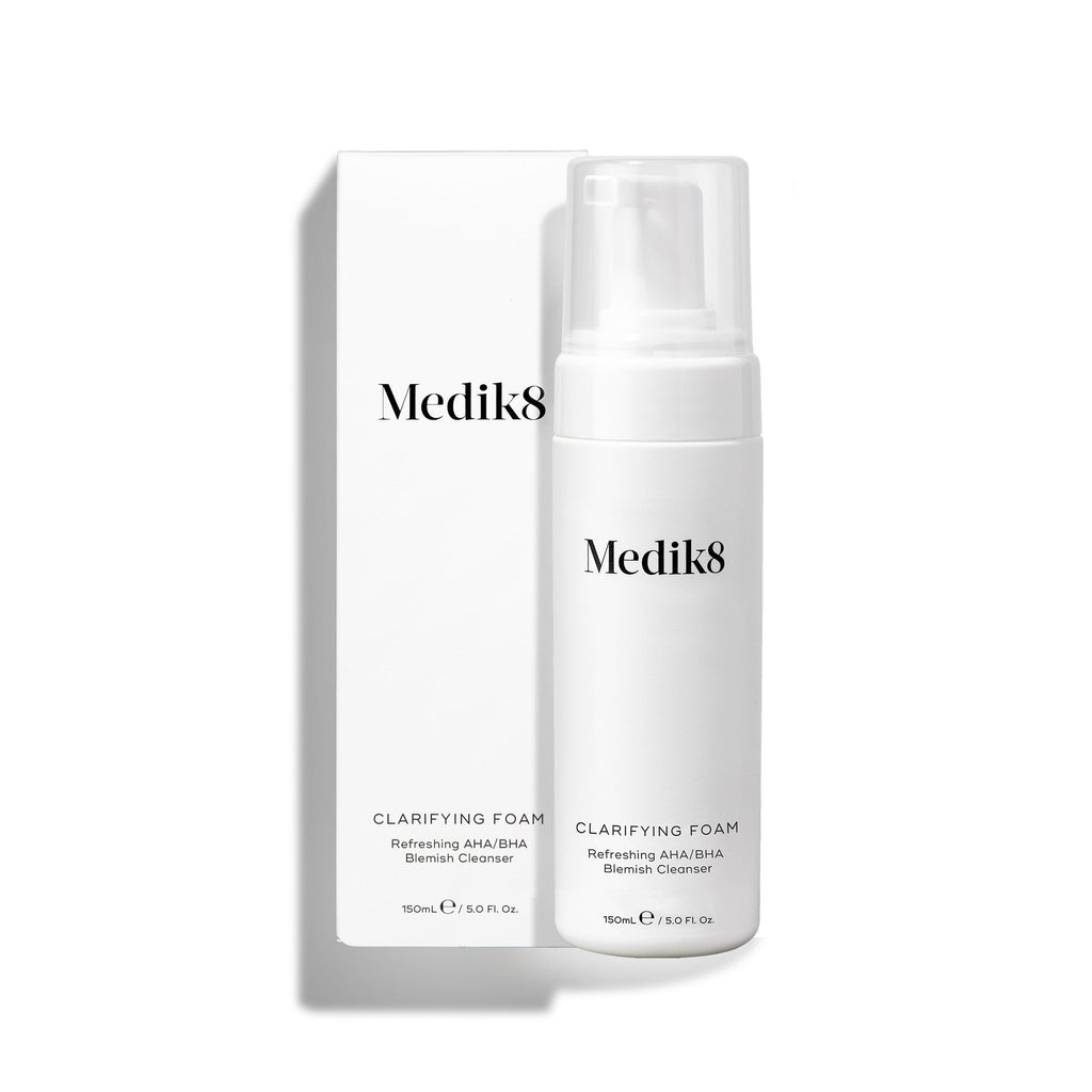 Fľaša s dávkovacím čerpadlom Medik8 Clarifying Foam stojí vedľa príslušného bieleho obalu na čistom bielom pozadí. Text na oboch označuje produkt ako osviežujúci čistiaci prípravok proti nedokonalostiam s obsahom AHA/BHA v objeme 150 ml.