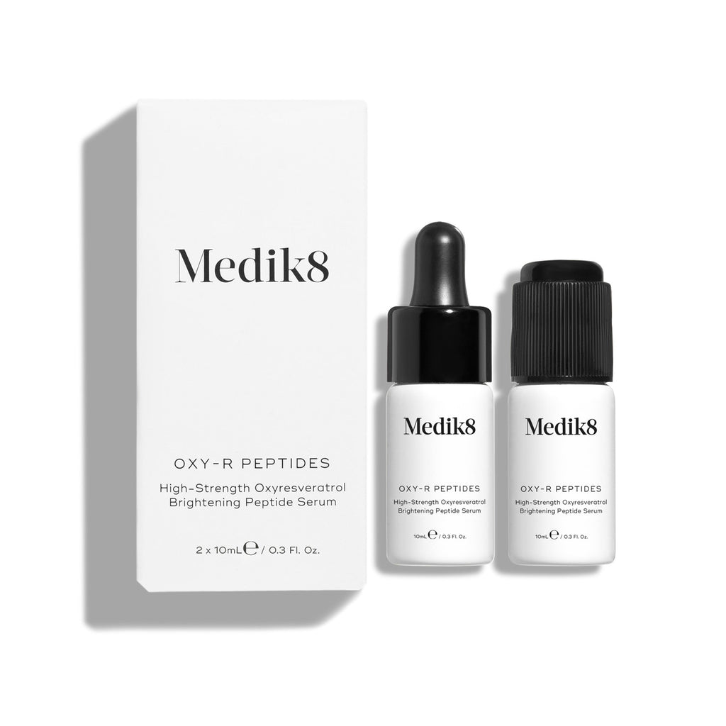 Na obrázku sú dve malé biele fľaštičky séra Oxy-R Peptides. Vedľa nich leží čierna kvapkačka a na bielom povrchu sú viditeľné dve kvapky žltého séra.