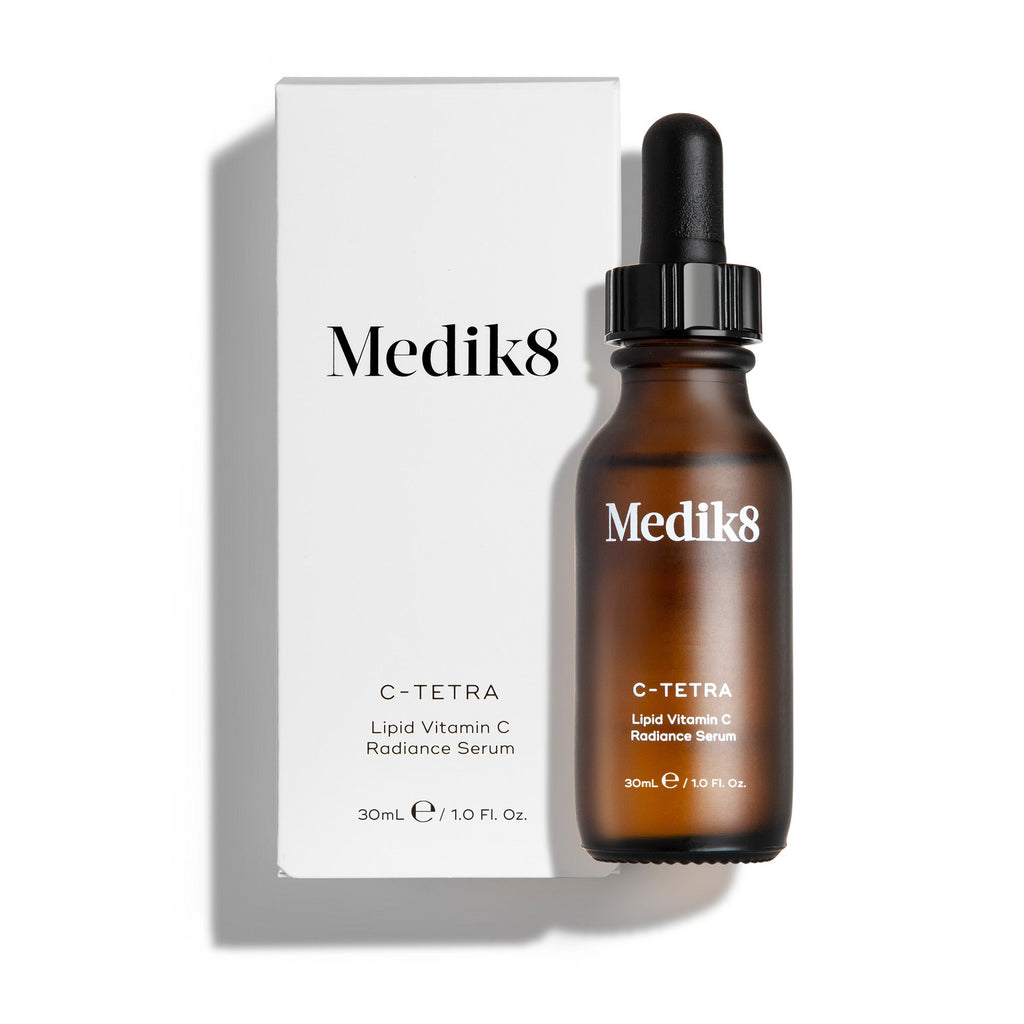 Fľaštička séra Medik8 C-Tetra Luxe a jej biela kartónová krabička sú zobrazené na čistom bielom pozadí s jemnými tieňmi. Na jantárovej sklenenej fľaštičke aj na bielej krabičke je umiestnené logo Medik8 a produkt je označený ako  C-TETRA LUXE Lipid Vitamin C Enhanced Radiance Serum  v balení s objemom 30 ml.