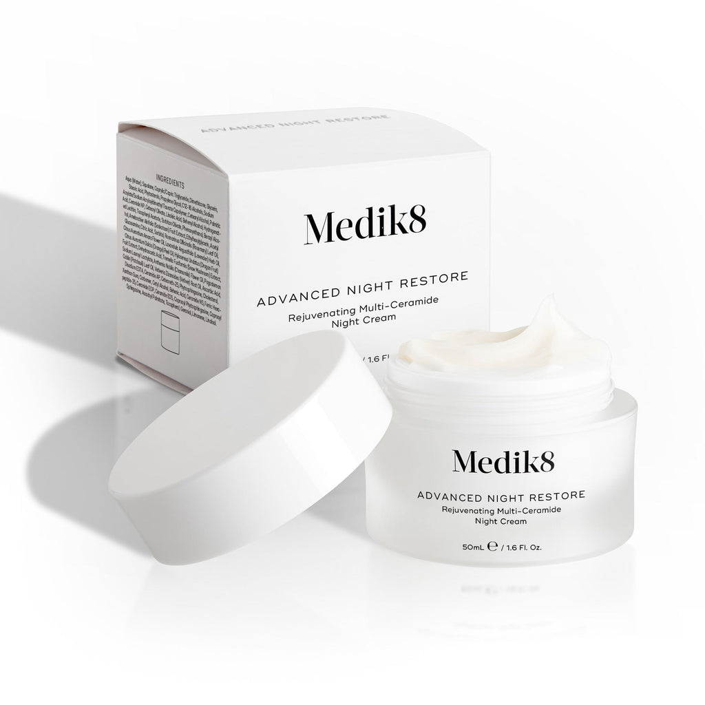 Kelímok krému Medik8 Advanced Night Restore je otvorený, jeho biele viečko sa oň opiera a za ním stojí krabička od produktu. Snímka zachytáva bohatú textúru krému a čistý dizajn balenia.