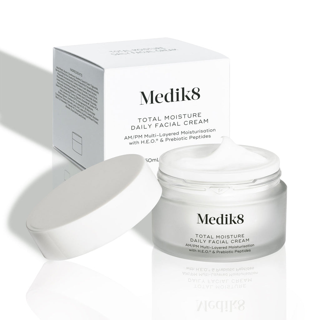 Matná nádobka s cestovným balením (12,5 ml) denného pleťového krému Medik8 Total Moisture stojí vedľa bieleho kartónového obalu. Výrobky sú vystavené na šedej štruktúrovanej ploche pred čistým bielym pozadím. Na oboch je nápis: „Viacvrstvová hydratácia na ráno a večer s H.E.O.™ a prebiotickými peptidmi.“