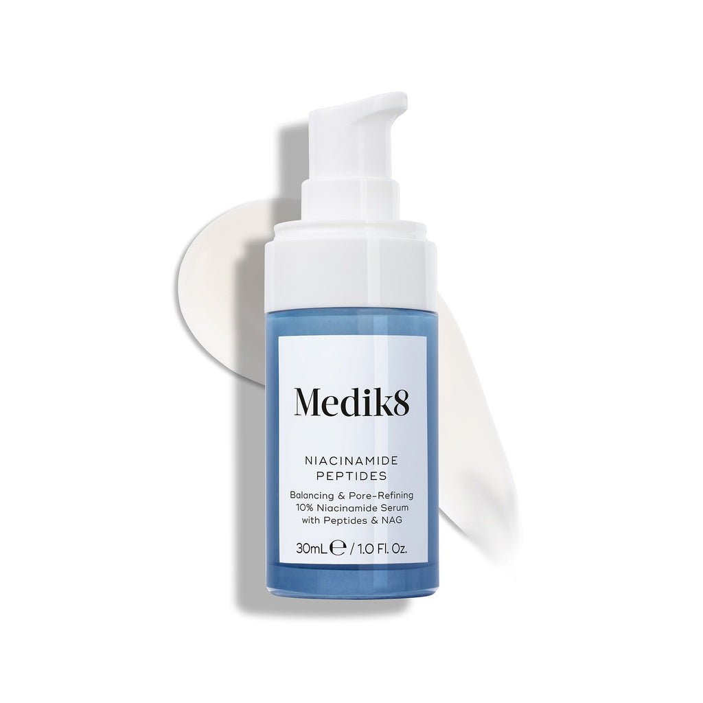 Fľaštička séra Medik8 Niacinamide Peptides s bielou pumpičkou leží na bielom podklade, pričom za ňou je viditeľná rozotretá vzorka bielej krémovej textúry tohto séra.