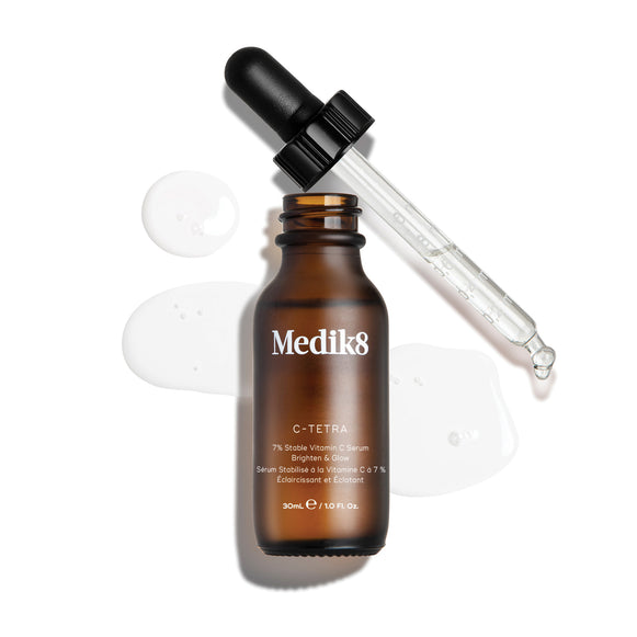 Horný pohľad na „packshot“ 30 ml fľaštičky séra Medik8 C-Tetra Lipid Vitamin C Radiance Serum. Jantárová sklenená fľaštička je otvorená a umiestnená v strede záberu, pričom jej čierne viečko s pipetou leží diagonálne za ňou. Číre tekuté sérum je umelecky rozliate a zhromaždené v kaluži na čistom bielom pozadí okolo fľaštičky, pričom na sklenenej pipete je viditeľných niekoľko kvapôčok. Jasné osvetlenie vytvára jemné tiene vľavo a dodáva obrázku klinický, no elegantný vzhľad.-15