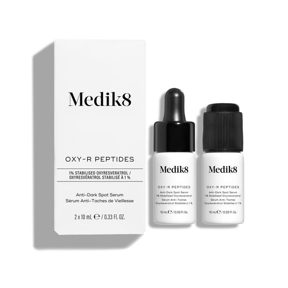 Biele produktové kartónové balenie séra Medik8 Oxy-R Peptides stojí vedľa dvoch malých bielych fľaštičiek séra. Jedna fľaštička má čiernu pipetu, druhá čierny uzáver.-34